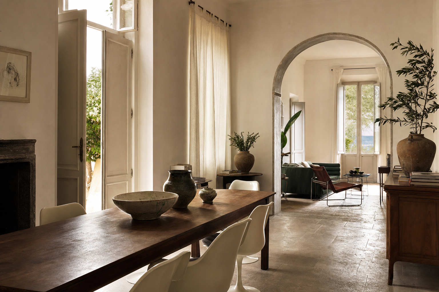 Een lichte woon-eetkamer met een houten tafel, moderne stoelen, keramieken schalen, linnen gordijnen en een verouderde stenen vloer bij hoge openslaande luiken.
