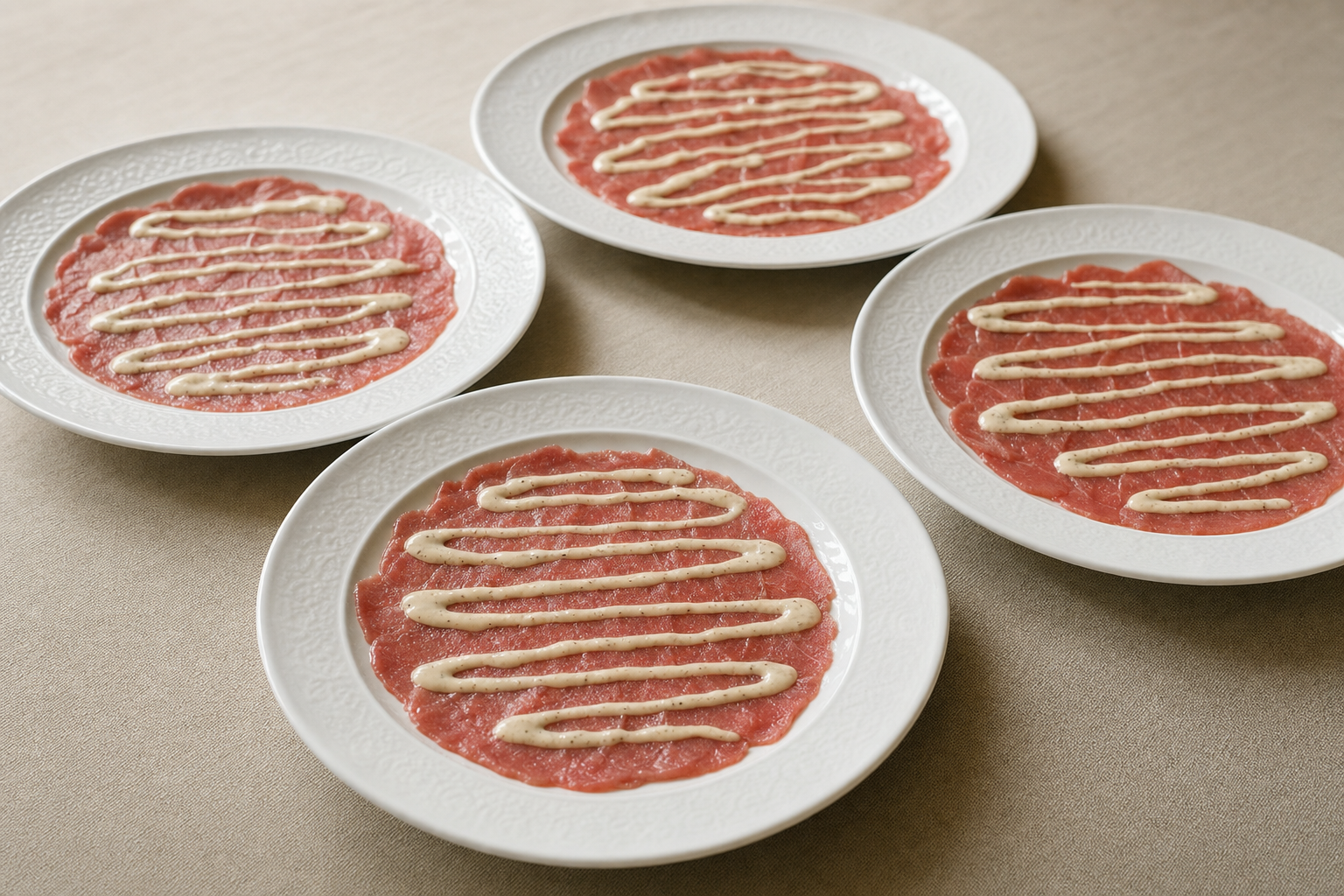 Vier witte borden met flinterdunne plakjes rauwe ossenhaas, waar dunne lijnen lichtbeige romige saus overheen zijn gedruppeld.